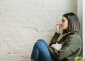 Side view of depressed young ethnic female millennial in warm sweater sitting leaning on wall and wiping tears with tissue at home