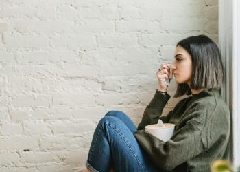 Side view of depressed young ethnic female millennial in warm sweater sitting leaning on wall and wiping tears with tissue at home