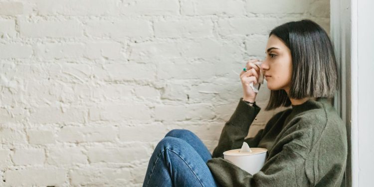 Side view of depressed young ethnic female millennial in warm sweater sitting leaning on wall and wiping tears with tissue at home